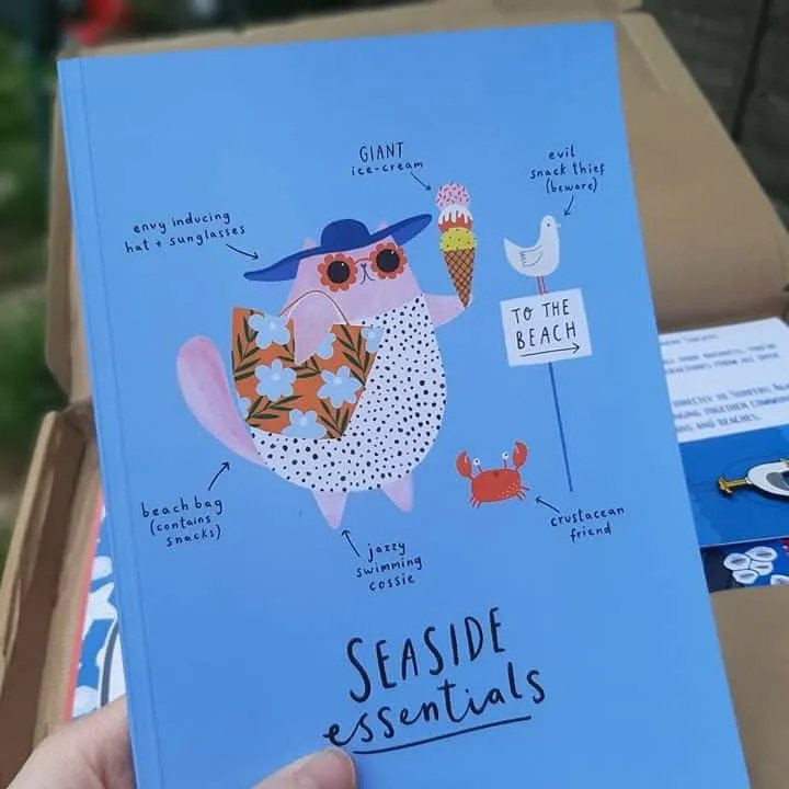 Ami is holding an A5 notebook, titled Seaside Essentials, created by Becky Hodgson.