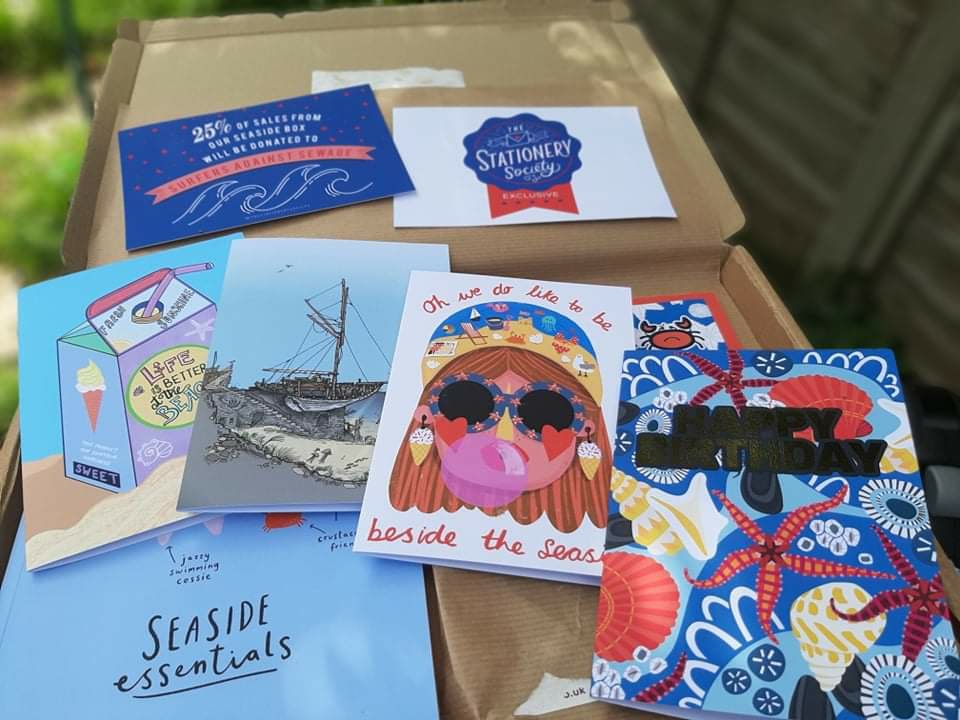 4 cards are placed, in a slight descending line, on top of the seaside-themed products. From left to right, the artists of the cards are Sofia Barton, Rho Rho Illustrations, Nichola Cowdery, and Lisa Kirkbride.